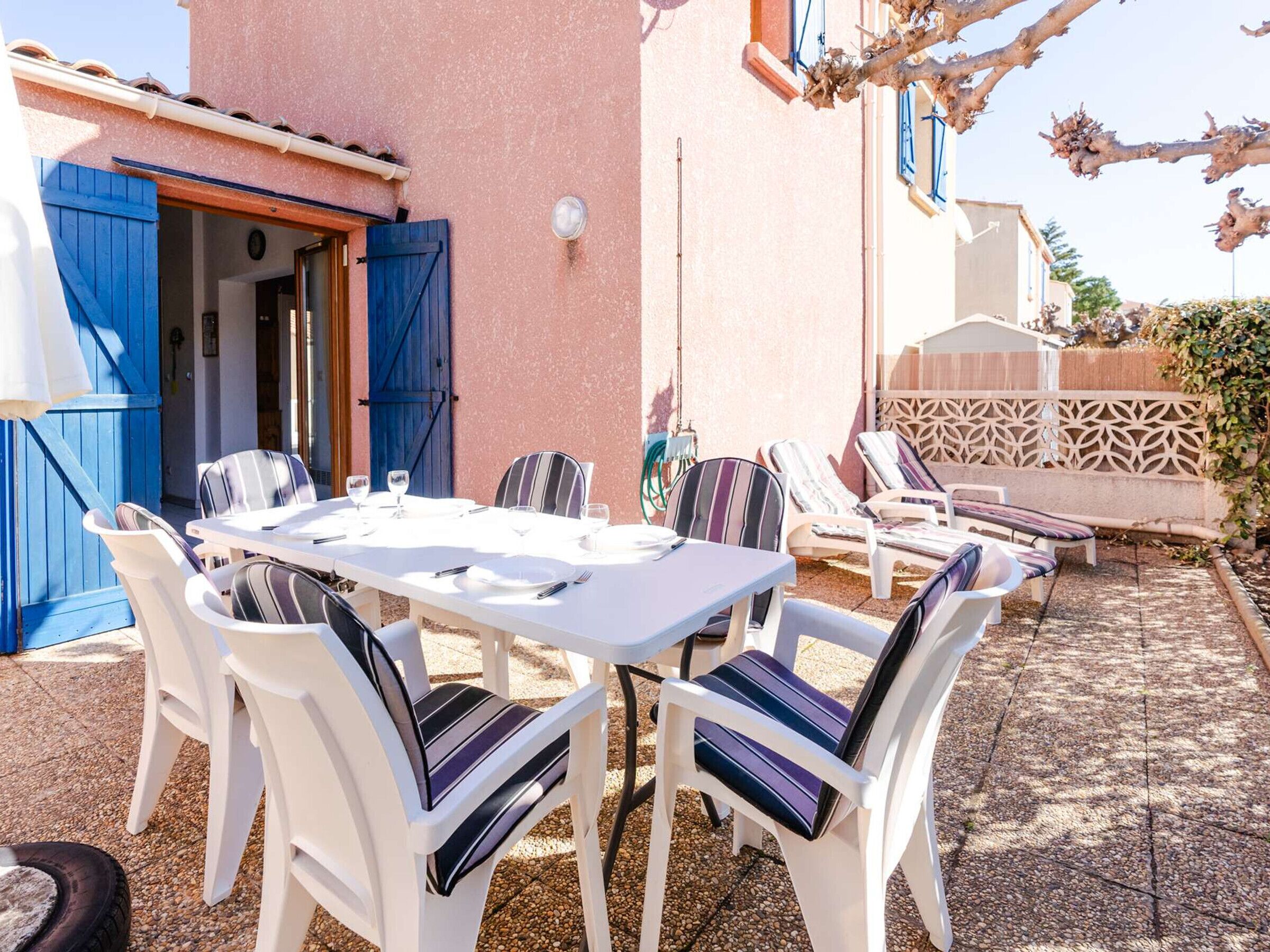 An outdoor patio with a white table set for dining, surrounded by white chairs with striped cushions.