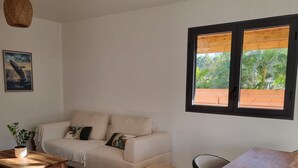 Ein Wohnzimmer mit Sofa, Couchtisch aus Holz, Esstisch und einem großen Fenster mit Blick ins Grüne.