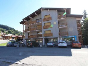 Ein mehrgeschossiges Gebäude mit Balkonen, ein Parkplatz mit mehreren Autos und eine Bergkulisse im Hintergrund.
