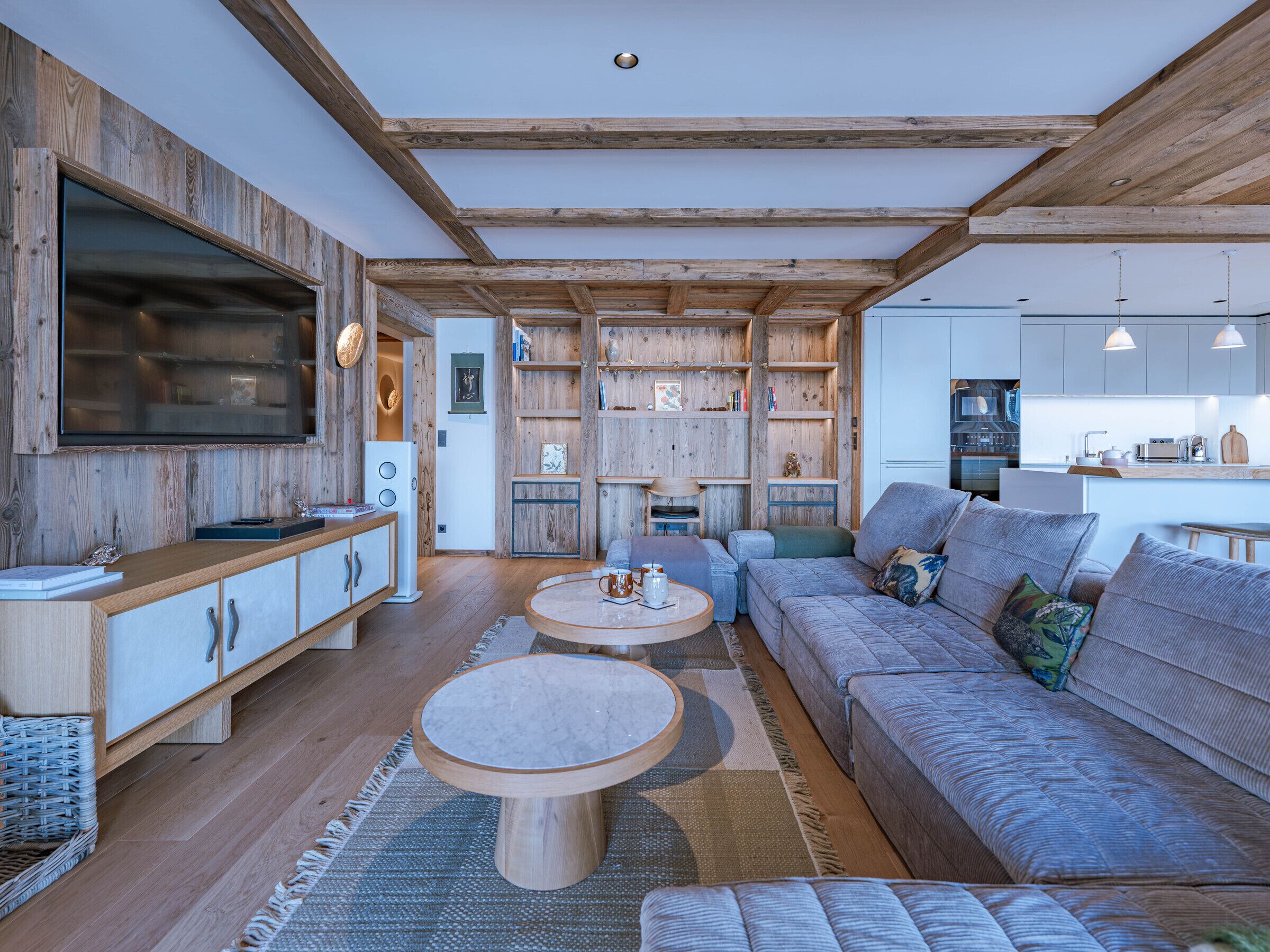 A modern living room with wooden beams, a built-in TV, a sofa, a coffee table, and a kitchen area.