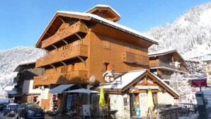 A wooden chalet-style building with balconies, a ski slope in the background, and a clear blue sky. - Charming apartment in village center - 4 guests (La Clusaz)