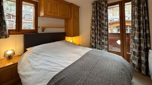 A bedroom with a bed, wooden cabinets, a hanging light fixture, and a view through a window. - Charming apartment in village center - 4 guests (La Clusaz)