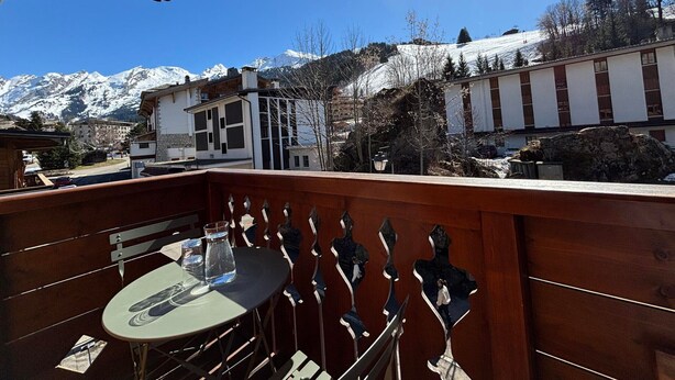 A balcony with a table and chairs, overlooking a snowy mountain and buildings. - Charming apartment in village center - 4 guests (La Clusaz)