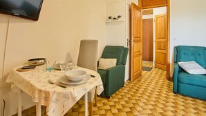 Ein Essbereich mit einem Tisch für zwei Personen, einem grünen Sessel, einem blaugrünen Sofa und einem an der Wand befestigten Fernseher.