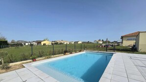 Una piscina rectangular con un borde empedrado, rodeada por una cerca y follaje.