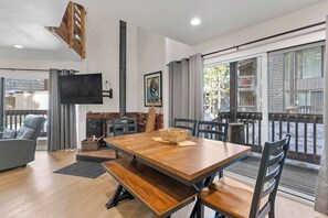 Ein modernes Wohnzimmer mit einem Holzesstisch, einer Couch, einem an der Wand montierten Fernseher und einem Balkon mit Blick ins Freie.