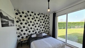 Une chambre avec un mur orné d’un motif géométrique, un lit avec une literie à rayures, une table de chevet, une lampe de lecture et une grande fenêtre donnant sur un jardin.