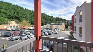 A parking lot with several cars, a building with a red facade, and a forested hillside in the background. - Studio, pets welcome, near city center (Lamalou-les-Bains)