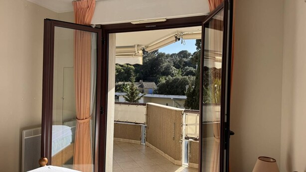 Quarto com cama, janela com cortinas, varanda com vista para edifícios e árvores, e luminária em uma mesa de cabeceira.