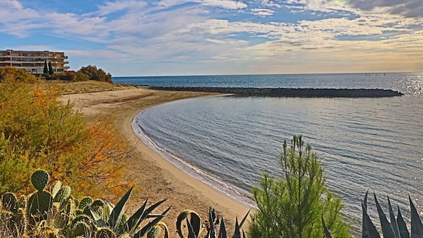 Eine Küstenlandschaft mit Sandstrand, einem Felsstollen und einem Gebäude mit Balkonen, das auf das Meer blickt.