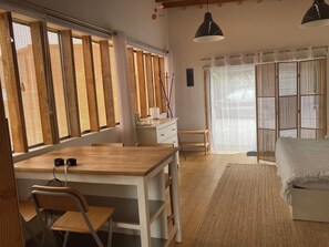 Une pièce avec un bureau en bois, des chaises, un lit et de grandes fenêtres équipées de stores.