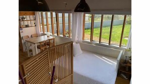 Una habitación con cama, mesa de comedor, sillas, cuna y ventanas amplias con vistas a follaje.