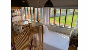 A room with a bed, a dining table, chairs, a crib, and large windows with a view of greenery. - L'atelier 800m from the sea, atypical accommodation in an old country neighborhood (Plouescat)