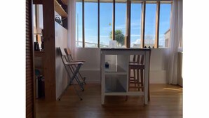 Una habitación con piso de madera, una pequeña mesa con sillas, un ventanal con persianas y un estante con objetos.