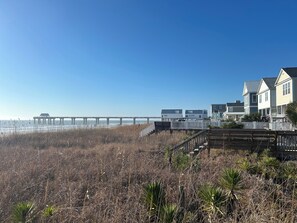 A coastal area with a pier extending into the water, a wooden boardwalk, and beachfront houses with a white fence.