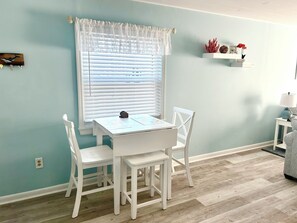 A dining area with a table and chairs, a window with blinds, a wall-mounted shelf with decorative items, and a lamp on a side table.