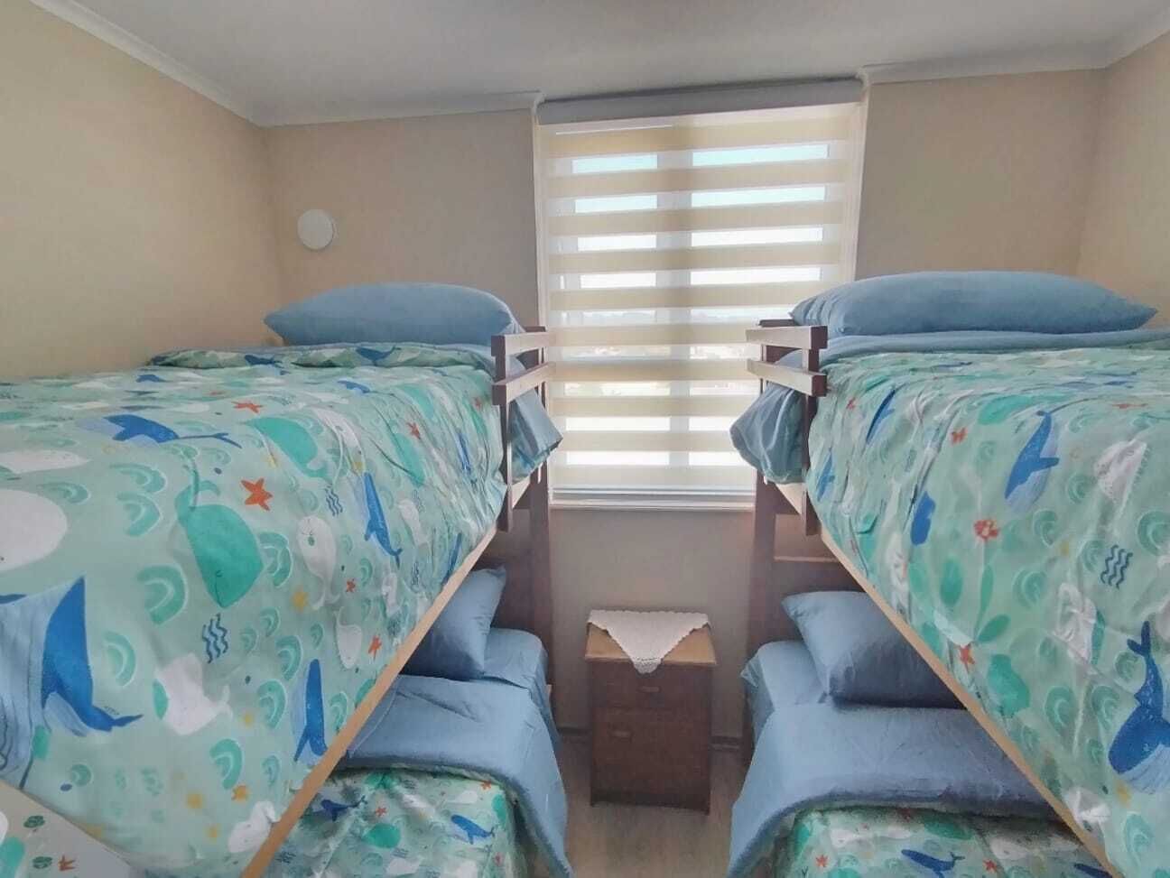 Een kamer met stapelbedden, een raam met zonwering, en een klein tafeltje met een witte tafelkleed.