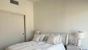 A bedroom with a bed, a nightstand, a lamp, a fur rug, and a vent on the wall. - Cozy 1-bedroom apartment in delightful Downtown Los Angeles with AC and gym (Los Angeles)