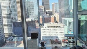 A view from a high-rise window overlooking a cityscape with modern buildings, a clear sky, and a window sill with sunglasses and a small decorative item. - Cozy 1-bedroom apartment in delightful Downtown Los Angeles with AC and gym (Los Angeles)