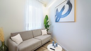 En stue med en beige sofa, et stort abstrakt maleri, en potteplante og et lille rundt bord med stearinlys og en fjernbetjening.