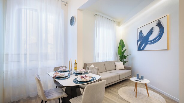 En moderne stue med et spisebord dækket til fire personer, en sofa, et lille sofabord og et stort abstrakt maleri på væggen.