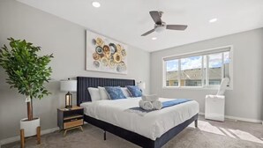 Un dormitorio con una cama, ventilador de techo, ventana, planta y una obra de arte en la pared.