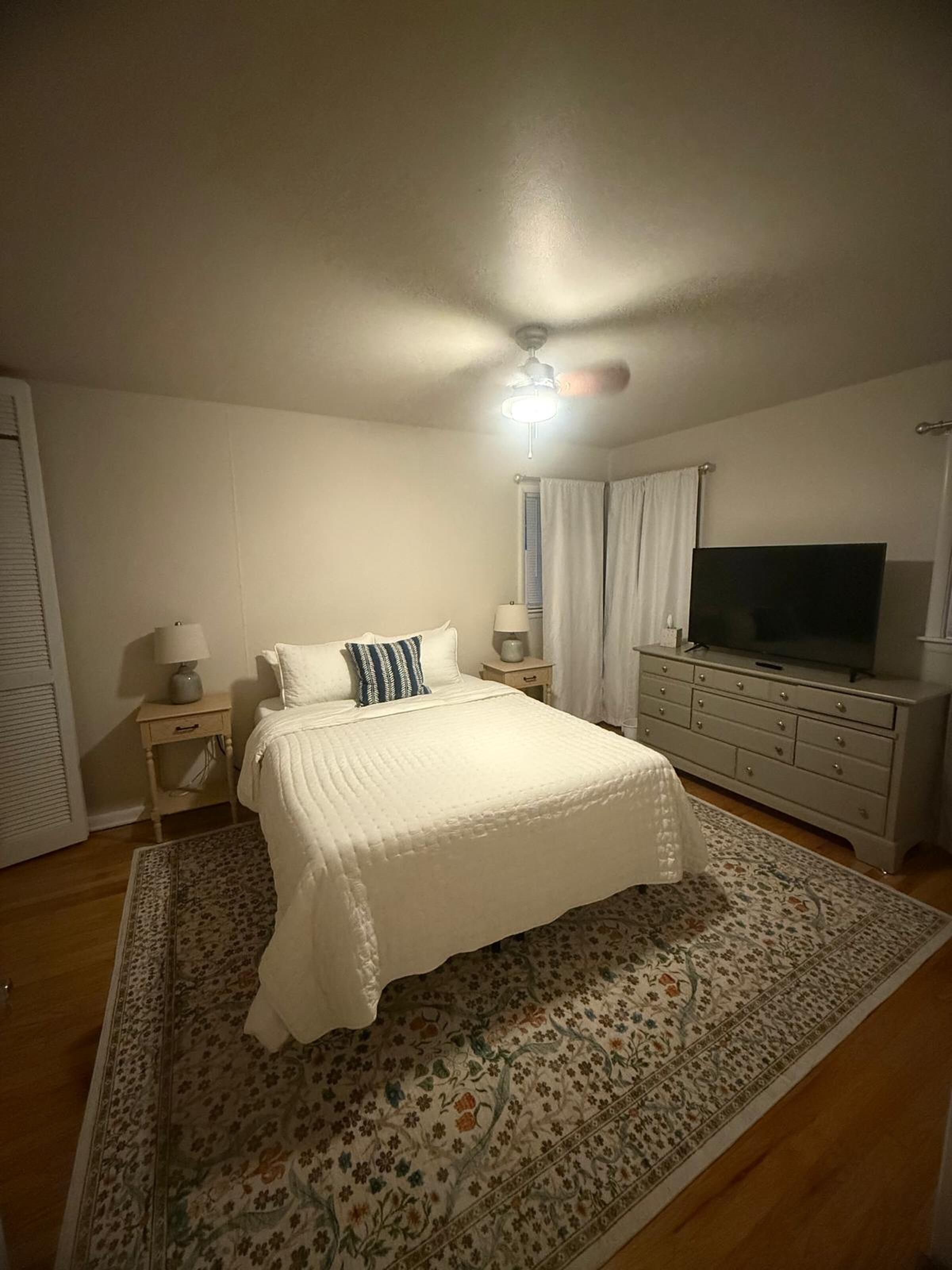 A bedroom with a bed, a TV, a ceiling fan, and a dresser.