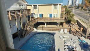 A pool area with a wooden deck, lounge chairs, and a view of a building with a blue roof. - Downtown Gulf Shores Beach -Best Spot in Complex W/Panoramic Views-Pet Friendly (Gulf Shores)