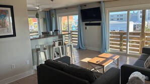 A living room with a sofa, a coffee table, a TV, and a balcony with a view of buildings. - Downtown Gulf Shores Beach -Best Spot in Complex W/Panoramic Views-Pet Friendly (Gulf Shores)
