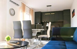 A modern living room with a blue sofa, a dining area with a table and chairs, and dark kitchen cabinets. - Amazing home in Dugobabe (Dugobabe)