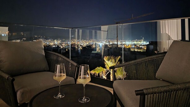 Ein Balkon mit zwei Stühlen und einem Tisch, zwei Weingläsern und Blick auf die beleuchtete Stadt.