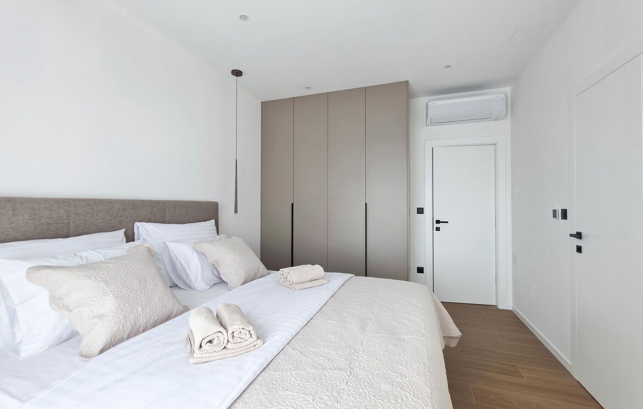 A bedroom with a bed, pillows, a bedside table, a wardrobe, and an air conditioning unit.