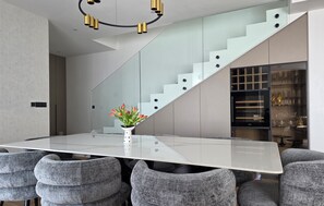 Ein moderner Essbereich mit einem Tisch mit Glasplatte, grauen Polstermöbelstühlen, einer Treppe mit Glastrennwand und einer dekorativen Vase mit Blumen.
