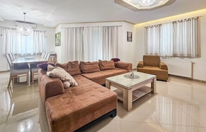 A modern living room with a sectional sofa, a coffee table, a dining area with a table and chairs, and large windows with curtains. - Awesome home in Donji Prolozac (Donji Prolozac)