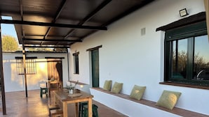 Un patio exterior con una mesa y sillas de madera, un banco integrado con cojines, y una ventana con vistas.