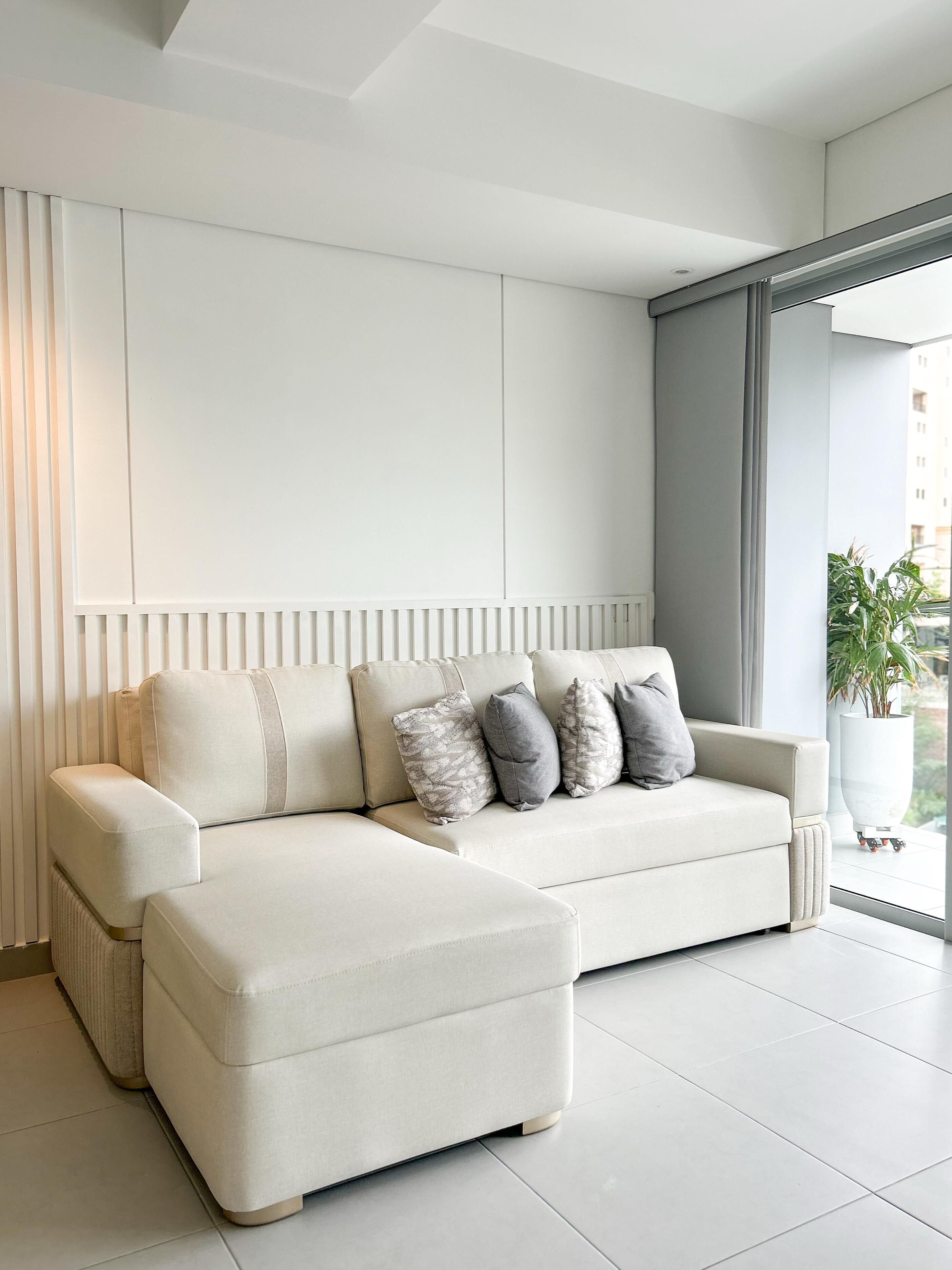 Ein modernes Wohnzimmer mit einem hellen Ecksofa, einer großen Topfpflanze und Blick ins Freie durch ein großes Fenster.
