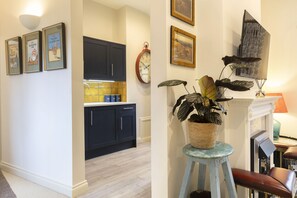 A hallway with a kitchen, framed pictures on the wall, a clock, and a potted plant.