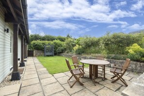 A patio with a round table and four chairs, a stone pathway, and a grassy area with plants and trees.