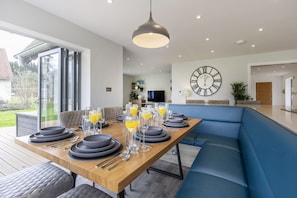 A modern dining area with a wooden table set for six, a large wall clock, and a blue bench. - Stunning Detached Countryside Home in Cheltenham (Cheltenham)