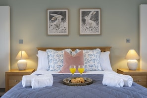 A neatly made bed with a quilted blanket, two bedside lamps, a tray with croissants and orange juice, and two framed artworks on the wall. - Stunning Detached Countryside Home in Cheltenham (Cheltenham)