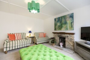 Een woonkamer met een groene bank, gestreepte fauteuils, een groen voetenbankje, een stenen open haard, een schilderij en een televisie.