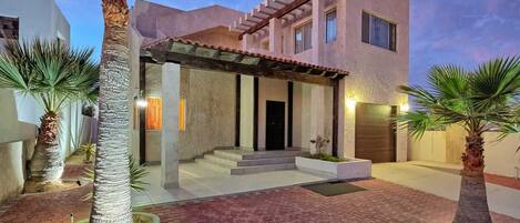 A modern two-story house with a covered entrance, palm trees, and a paved driveway.