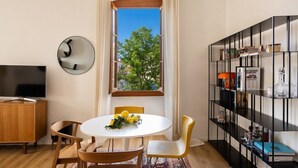 A modern living room with a round table, chairs, a TV, a bookshelf, and a window with a view of trees. - Casa Bianca – Charme & vue mer au cœur d’Erbalunga (Brando)