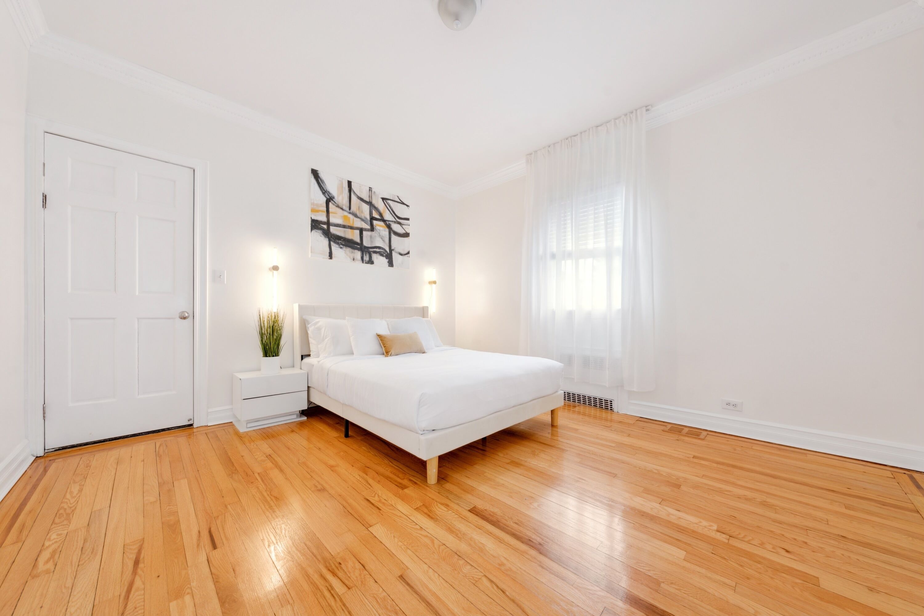 A bedroom with a wooden floor, a white bed, a nightstand with a plant, a wall-mounted artwork, and a white door.
