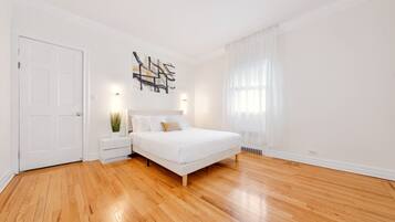 A bedroom with a wooden floor, a white bed, a nightstand with a plant, a wall-mounted artwork, and a white door.