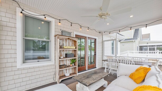 Un patio techado con un muro de ladrillo blanco, un ventilador de techo, luces colgantes, un banco de madera, mobiliario blanco y una valla blanca al fondo.