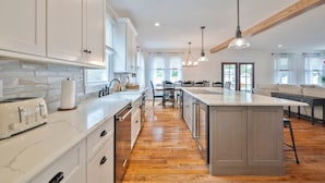 Una cocina moderna con gabinetes blancos, encimera de mármol, suelo de madera e iluminación colgante.