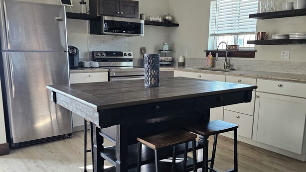 A modern kitchen with a central island, stainless steel appliances, and a window with blinds. - Pleasant 1-bedroom cottage in charming Manhattan (Manhattan)