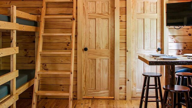 A wooden interior with a bunk bed, a dining table with stools, and a TV mounted on the wall. - Family Cabin in the heart of The Pennsylvania Grand Canyon (Wellsboro)