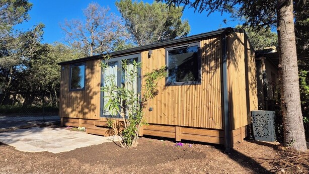 Una cabaña de madera con ventanas amplias, un pequeño jardín y un árbol cercano.